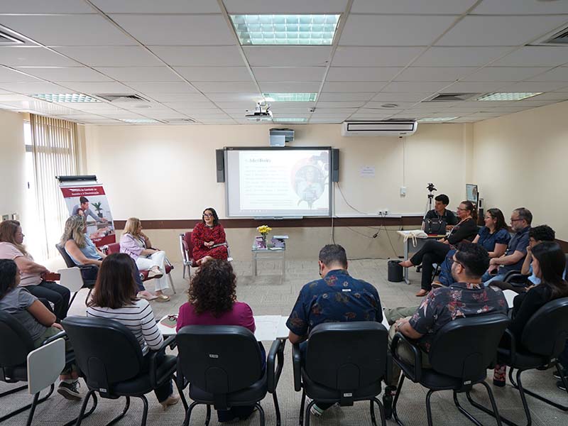 Uma sala de aula clara, com cerca de 20 pessoas sentadas em cadeiras dispostas em semicírculo. No centro, uma mulher de vestido vermelho fala enquanto outra mulher de blusa rosa claro e calça branca escuta. Ao fundo, uma tela projeta uma imagem com texto.