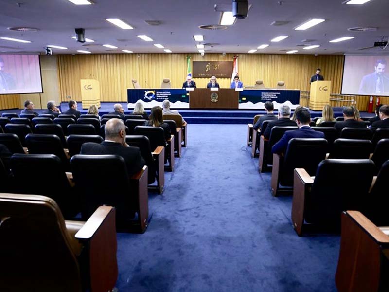 Esta é uma fotografia em plano médio, tirada de frente, mostrando uma sala de audiência com um piso azul e cadeiras escuras. No centro, uma mesa comprida de madeira está disposta de forma horizontal. Três pessoas, em trajes formais, estão sentadas atrás da mesa, de frente para um auditório com poucas pessoas.