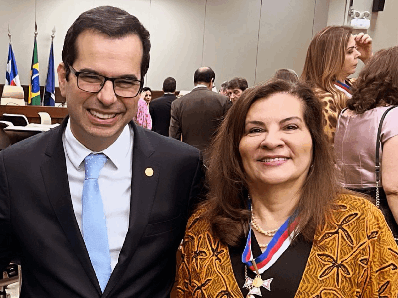 Um homem e uma mulher sorriem para a câmera, posando lado a lado. O homem, de terno escuro, gravata azul e óculos, está à esquerda da imagem. A mulher, à direita, usa um casaco marrom e preto e ostenta uma medalha no pescoço. O cenário parece ser um plenário ou auditório, com bandeiras e pessoas ao fundo.