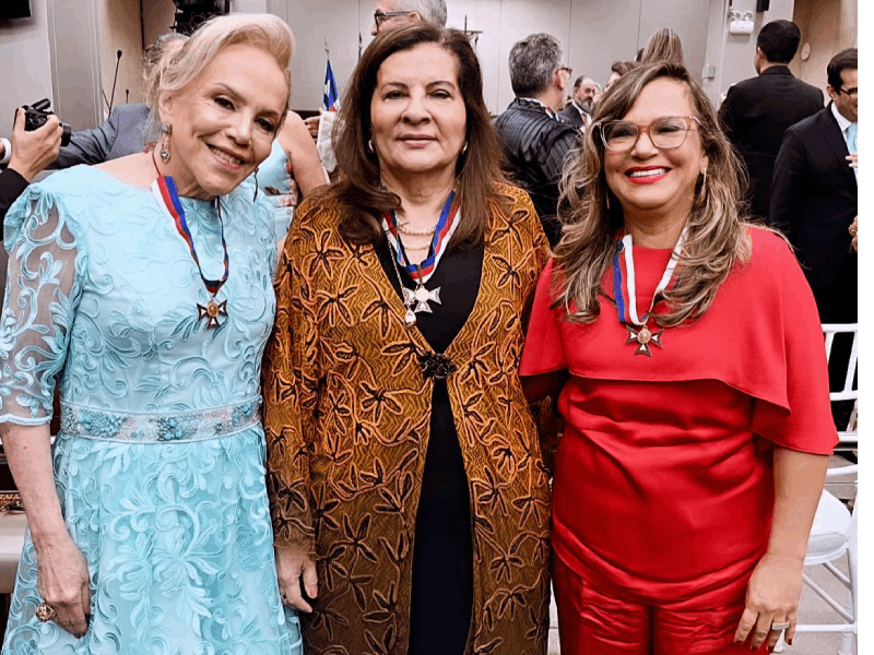 Três mulheres posam sorrindo. A do centro, de casaco marrom e preto, tem uma medalha no pescoço. Ela está ladeada por duas amigas, uma de vestido azul e outra de blusa vermelha, ambas também condecoradas com medalhas. Ao fundo, um auditório movimentado.