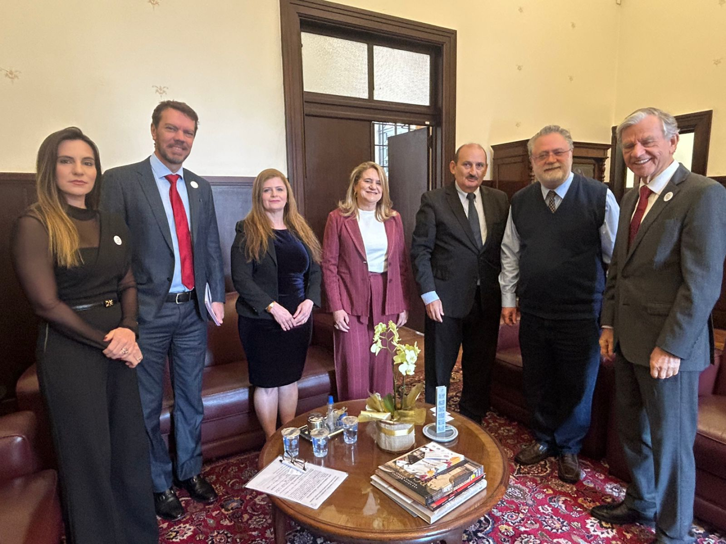 Uma fotografia em que seis pessoas, cinco homens e uma mulher, posam para uma foto em uma sala de estar. No centro da sala há uma mesa de centro de madeira, cheia de livros e copos.