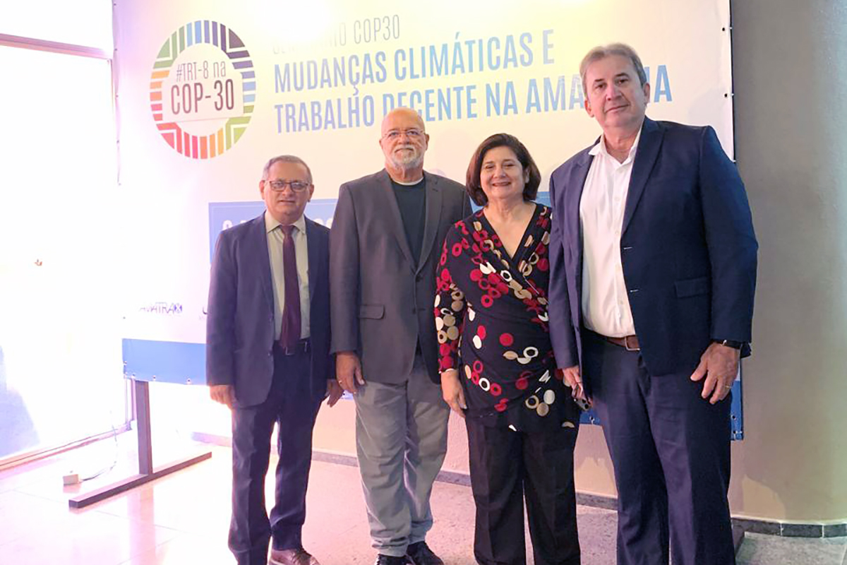 Fotografia de quatro pessoas, sendo três homens e uma mulher, em pé e lado a lado. Eles estão em um evento, com um banner ao fundo sobre "Mudanças Climáticas e Trabalho Decente na Amazônia", com o logotipo "#TRT-8 na COP-30". As pessoas vestem trajes formais e sorriem discretamente para a câmera.