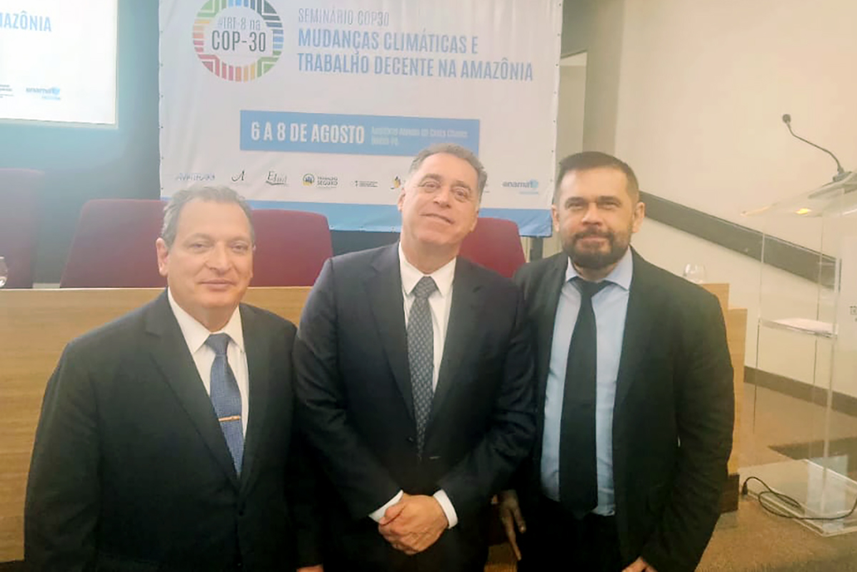 Três homens de terno e gravata estão em pé, em um auditório com cadeiras vermelhas e um púlpito ao lado. Ao fundo, um banner do "Seminário COP30" sobre mudanças climáticas é visível. O homem do centro sorri, enquanto os outros dois olham seriamente para a foto.