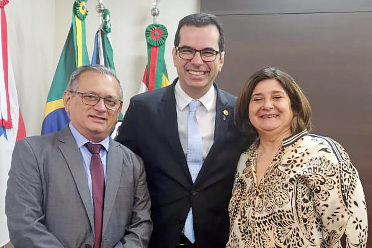 Em um ambiente interno com bandeiras ao fundo, três pessoas posam para uma foto. No centro, um homem alto de óculos e terno sorri abertamente. Ele é ladeado por um homem de terno cinza à sua direita e uma mulher de cabelos curtos e blusa estampada à sua esquerda.