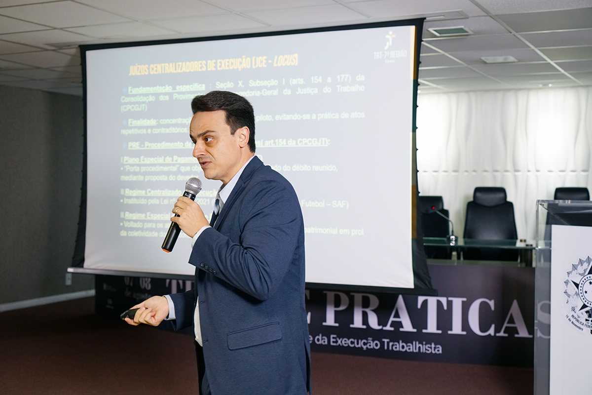 Em um auditório, um homem de terno azul escuro fala ao microfone, que segura com a mão direita. Com a esquerda, ele segura um passador de slides. Ao fundo, uma tela de projeção exibe um texto em tópicos.