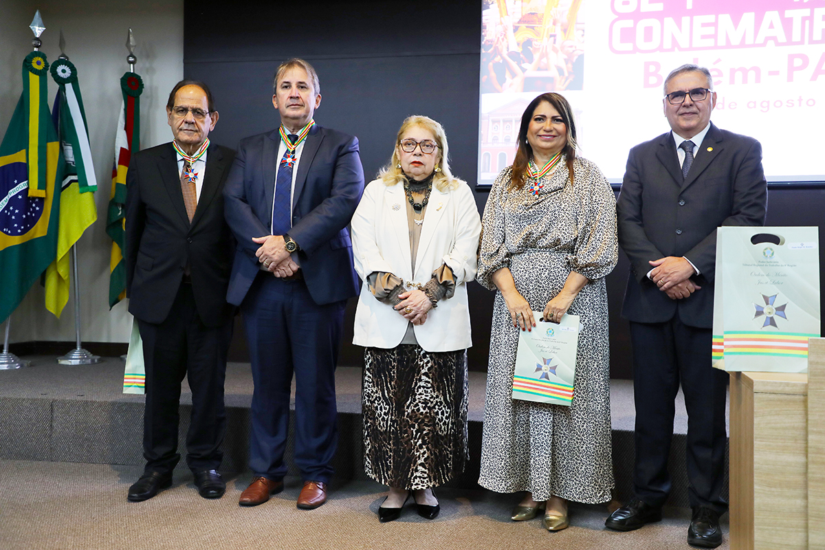 Fotografia de cinco pessoas posando para foto em um evento solene. Em um palco, três homens de terno e duas mulheres com vestidos formais estão em pé, lado a lado. Quatro deles usam medalhas de honra penduradas no pescoço. À esquerda, há bandeiras do Brasil e de outra instituição. Ao fundo, um telão e, à direita, um púlpito com o que parecem ser certificados, indicando uma cerimônia de outorga.