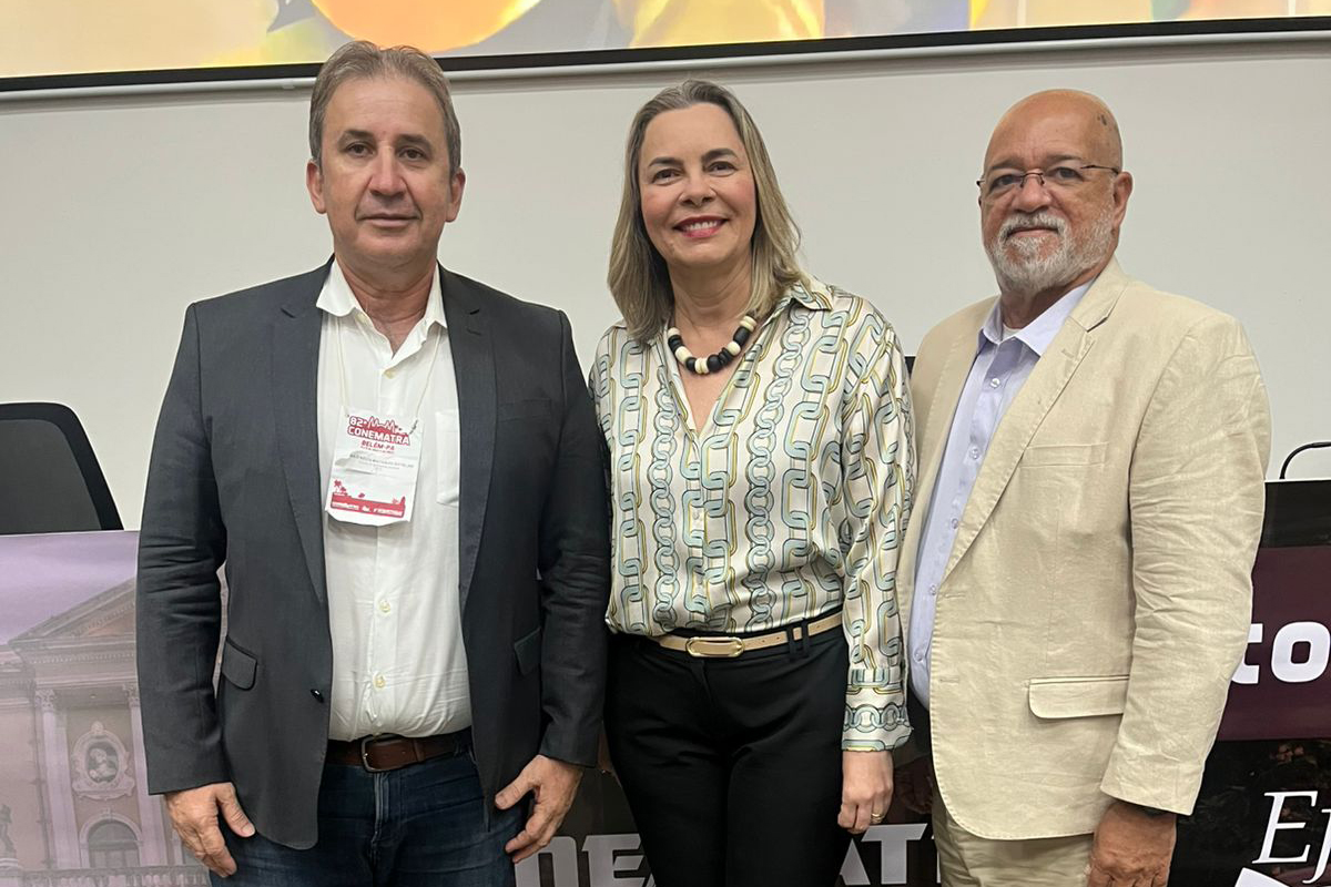 Fotografia de três pessoas posando juntas em um evento profissional. Uma mulher loira e sorridente está no centro, vestindo uma blusa estampada, entre dois homens. O homem à sua esquerda usa um blazer escuro e um crachá de evento. O homem à sua direita, de barba e óculos, veste um blazer de cor clara. Ao fundo, um painel desfocado sugere que eles estão em um congresso ou seminário.