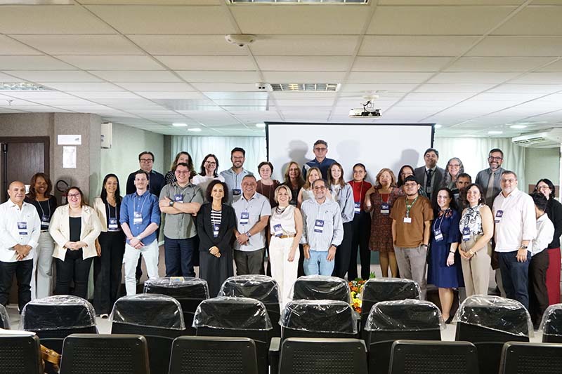Uma fotografia em ambiente fechado mostra um grupo de cerca de 30 pessoas, a maioria sorrindo e olhando para a câmera, de pé em três filas. Ao fundo, uma tela de projeção em branco e cadeiras vazias em primeiro plano.