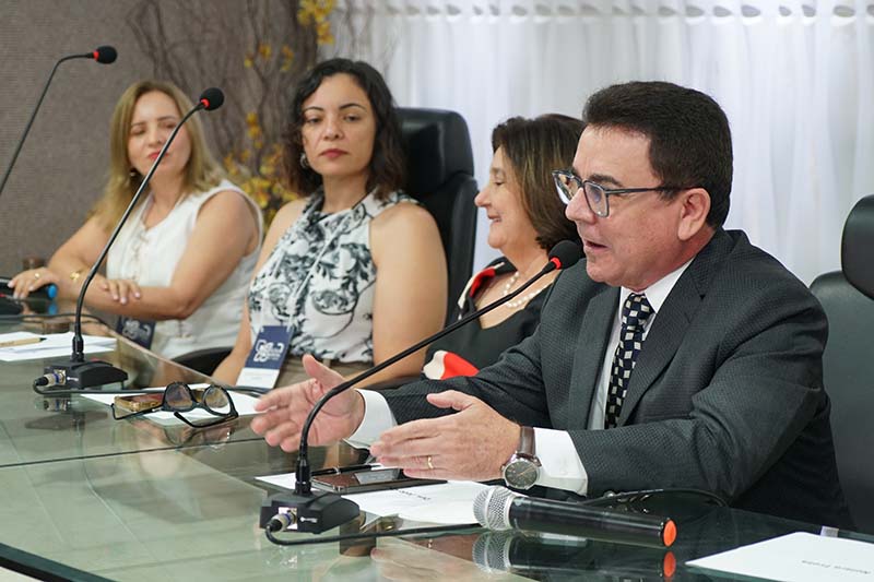 Uma fotografia em close mostra um homem de terno falando ao microfone, com as mãos gesticulando. Atrás dele, três mulheres o observam atentamente. A mesa é de vidro e à frente de cada participante há um microfone, óculos e alguns papéis.