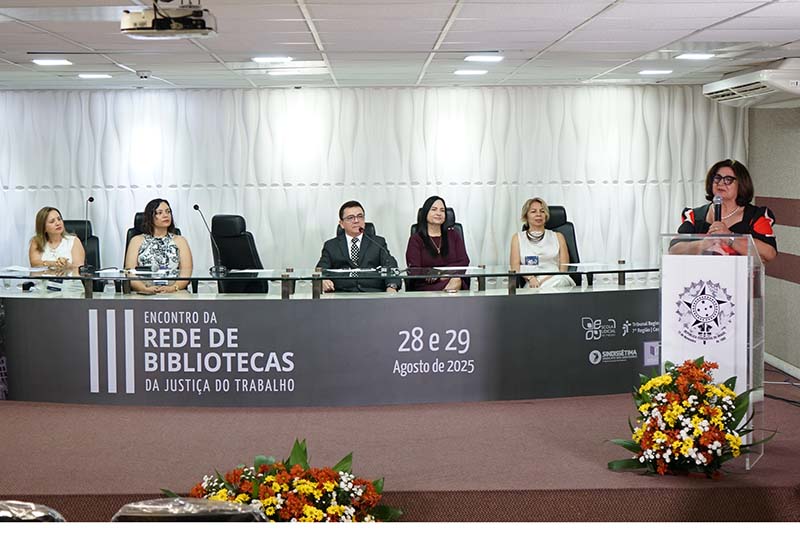 Uma fotografia mostra seis pessoas em uma mesa semicircular em um auditório. A mulher da extrema direita, de vestido estampado, está de pé em um púlpito, com uma das mãos sobre ele e a outra gesticulando. As outras cinco pessoas estão sentadas, com as mãos sobre a mesa, voltadas para a mulher