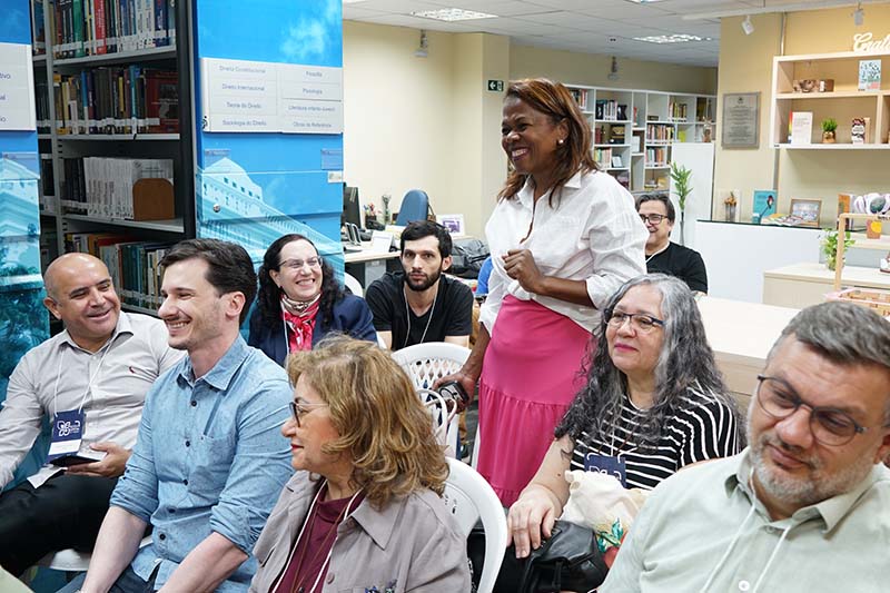 Uma mulher de blusa branca e saia rosa, de pé no centro de uma sala, sorri enquanto olha para a direita. Ao seu redor, as outras pessoas sentadas em cadeiras brancas a observam. Ao fundo, uma estante de livros e uma mesa com objetos de decoração.