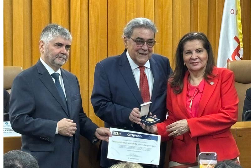 Três pessoas estão de pé em uma sala de reuniões. Um homem, à esquerda, e uma mulher, à direita, seguram as pontas de um certificado. Ao centro, outro homem com óculos e gravata segura uma medalha dourada em um estojo.