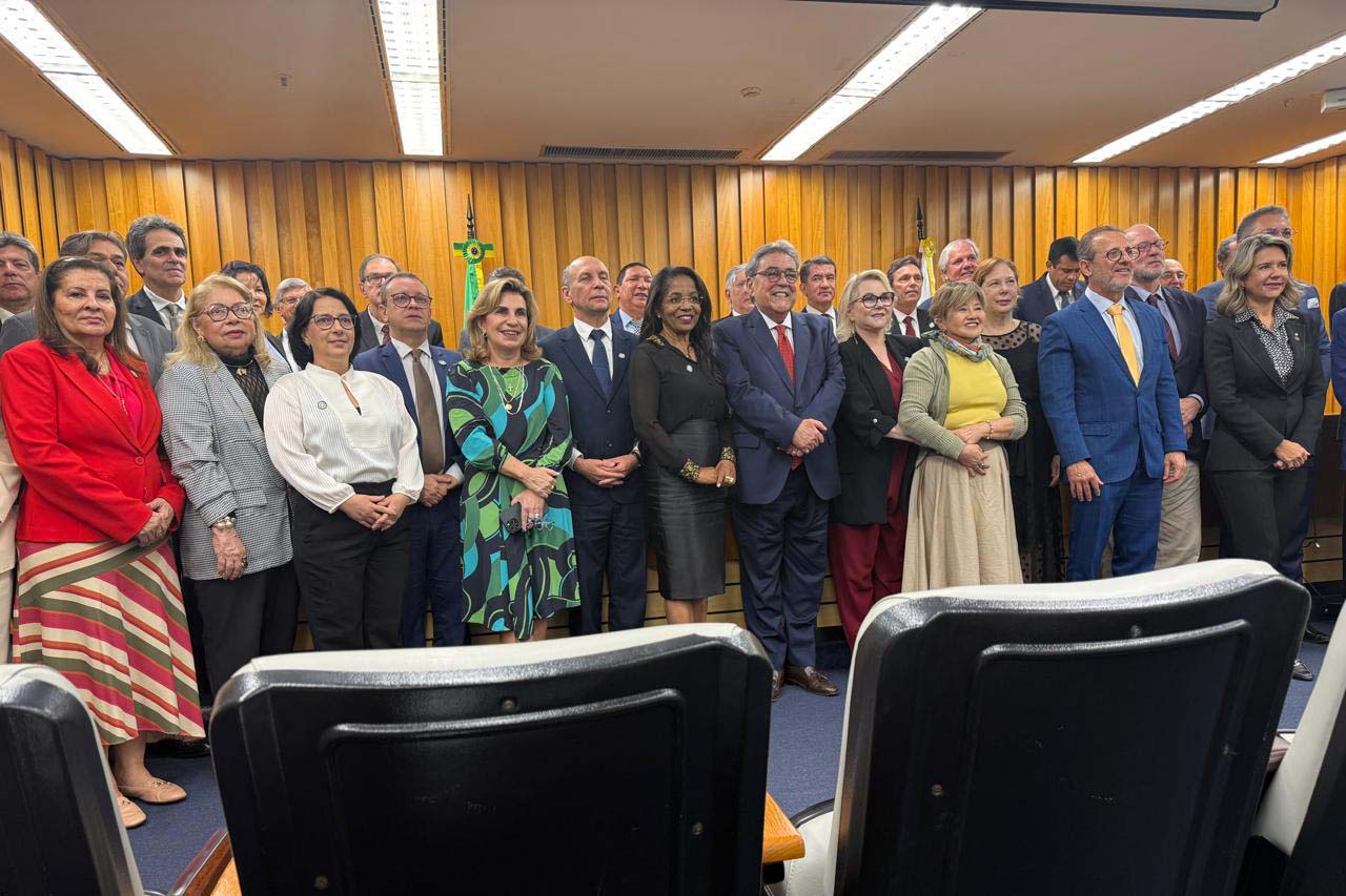 Um grupo de cerca de 20 pessoas, entre homens e mulheres, está de pé em uma sala de madeira com carpetes azuis, formando duas fileiras. Estão todos sorrindo, e alguns usam óculos e gravatas. A imagem é vista de uma perspectiva mais baixa, com cadeiras de escritório em primeiro plano.