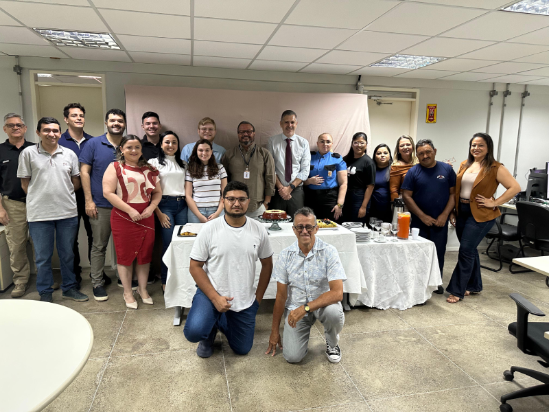Um grupo diversificado de 17 pessoas, incluindo homens e mulheres de várias idades, está reunido em uma sala de escritório para uma celebração. Ao centro, há uma mesa com um bolo e bebidas, e a maioria das pessoas está sorrindo, indicando um ambiente festivo e acolhedor.