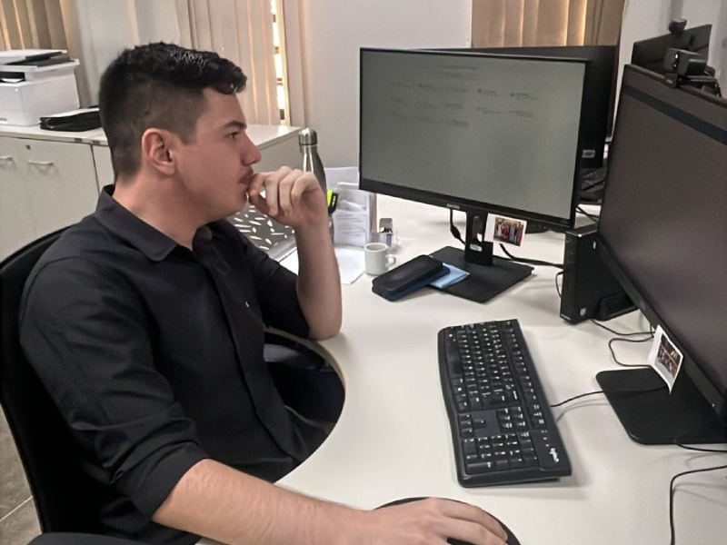  Um homem jovem de perfil, com cabelo preto e curto, está sentado em uma cadeira de escritório. Ele está concentrado, com a mão no queixo, olhando fixamente para um monitor de computador. Vestindo uma camisa social preta, ele tem a outra mão sobre o mouse, e o teclado está logo à frente, indicando um momento de reflexão ou trabalho intenso.
