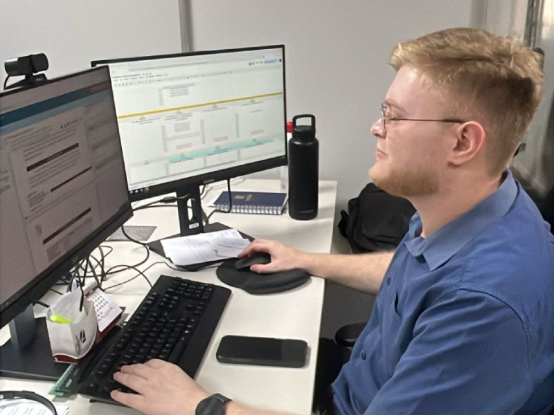 Um homem jovem, de cabelo loiro-avermelhado e óculos, está sentado de perfil em uma mesa de escritório, concentrado em dois monitores de computador. Ele usa uma camisa azul e suas mãos estão sobre o teclado e o mouse, indicando que está trabalhando. Uma garrafa térmica preta e alguns papéis estão visíveis na mesa, complementando o ambiente de trabalho.