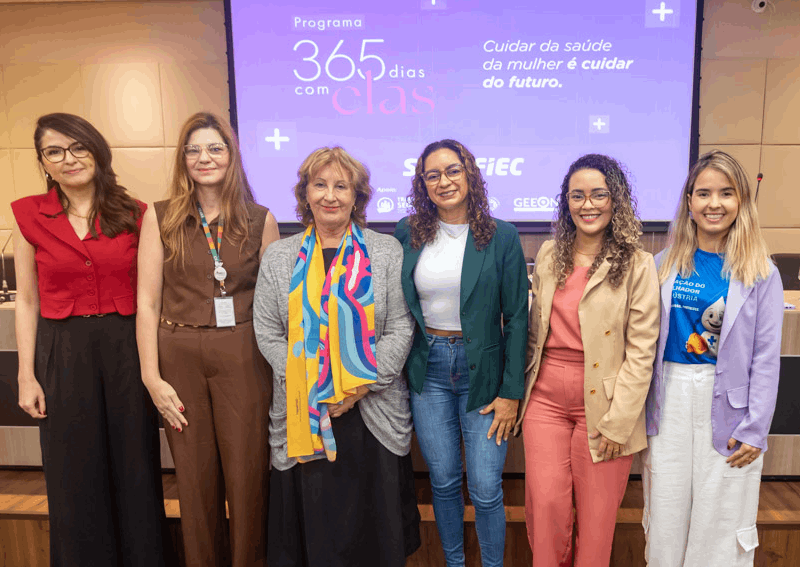 Uma fotografia mostra sete mulheres sorrindo para a câmera, em pé, em frente a uma tela de projeção com o texto "Programa 365 Dias com Elas - Cuidar da saúde da mulher é cuidar do futuro". A iluminação clara do ambiente realça os detalhes de suas roupas e penteados, com algumas usando blazers, camisas e calças, e outras exibindo acessórios coloridos como um lenço estampado. No lado direito da tela de projeção, há o logo da FIEC, do SESI e do GEEON.