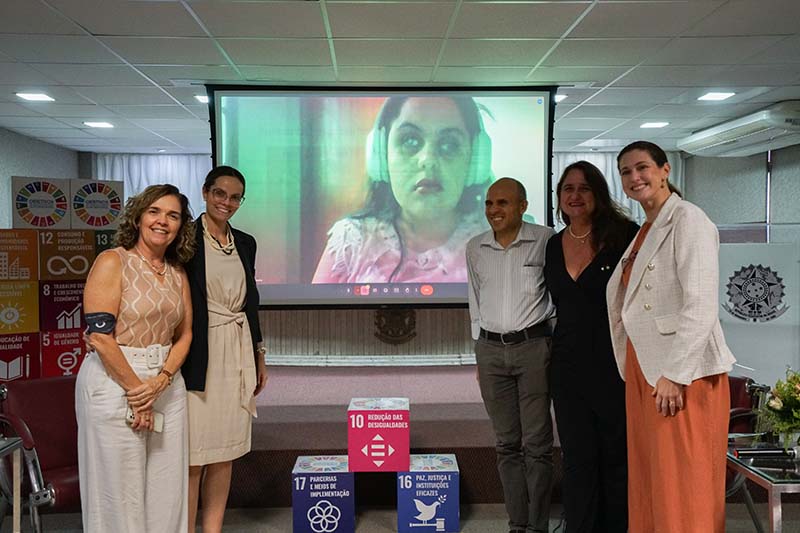  Cinco pessoas, quatro mulheres e um homem, estão alinhadas no palco de um auditório. Ao fundo, um telão mostra uma mulher em videoconferência. À frente do palco, há três cubos com os logotipos dos Objetivos de Desenvolvimento Sustentável (ODS) da ONU.