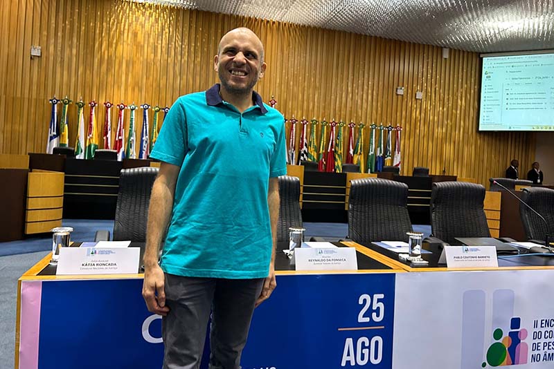  Um homem careca com barba e camisa polo azul-turquesa sorri para a câmera. Ele está de pé em um grande salão com bandeiras ao fundo. Na mesa à sua frente, há placas de identificação com nomes e um banner no chão faz referência a um evento.