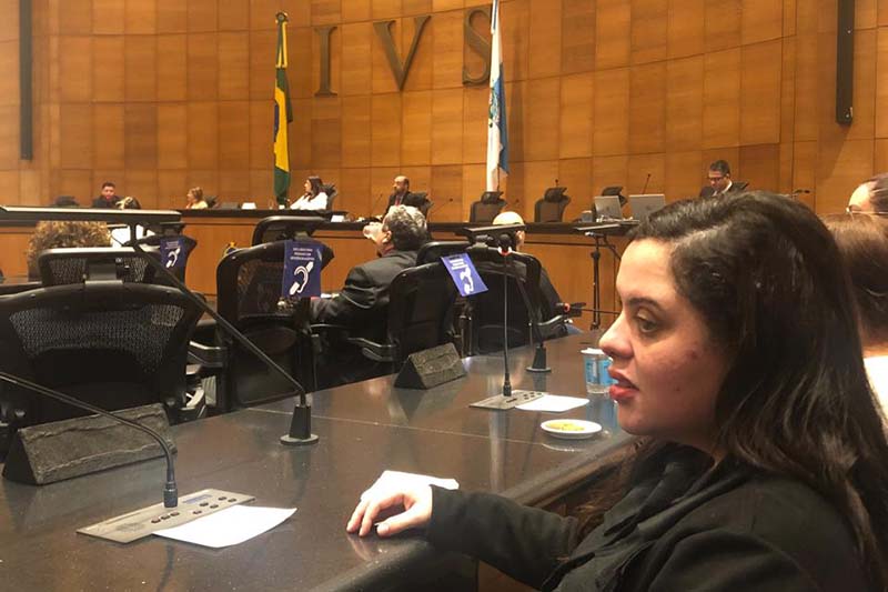  Uma mulher de perfil, com jaqueta preta, está sentada em uma mesa de audiências. Ela observa atentamente a frente, com o braço apoiado na mesa. Ao fundo, outras pessoas estão sentadas e o novo Símbolo Internacional de Acessibilidade está exposto em pequenos banners sobre a mesa.