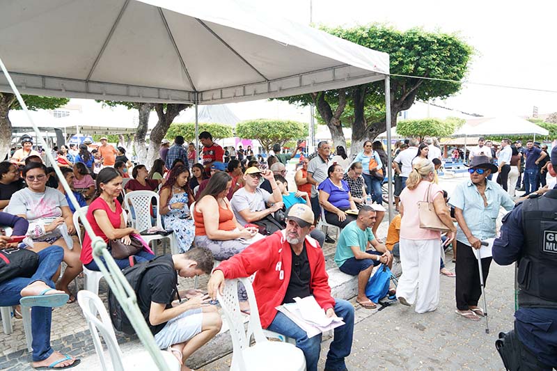 Uma imagem mostra uma área externa sob uma tenda branca, onde várias pessoas de diferentes idades estão sentadas em cadeiras brancas e de plástico. Ao fundo, mais pessoas circulam, e há vegetação e edifícios à distância. Um guarda municipal está visível no canto inferior direito.