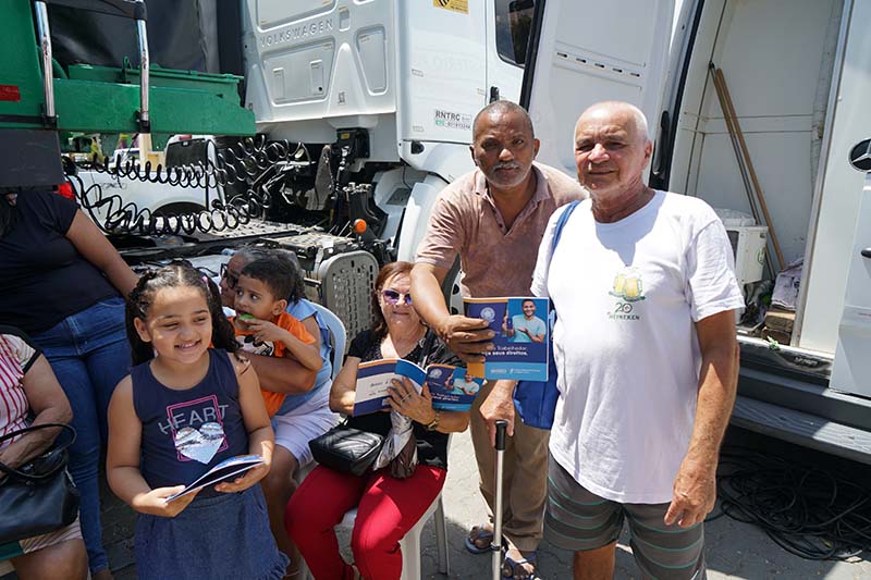 Uma foto mostra um grupo de pessoas de diferentes idades posando em frente à lateral de um grande caminhão branco. A frente do grupo, uma menina sorridente segura uma cartilha. Três adultos, incluindo um homem de bigode e um idoso, também seguram cartilhas.