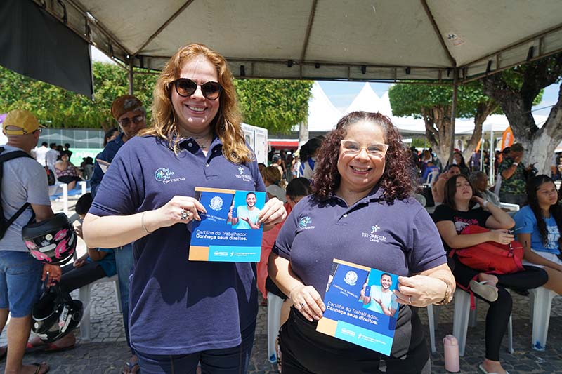 Uma cena mostra uma área externa sob uma grande tenda branca. Várias pessoas de diversas idades estão sentadas em cadeiras de plástico brancas. Ao fundo, outras pessoas circulam, e é possível ver árvores e prédios distantes. No canto inferior direito, um guarda municipal está visível.