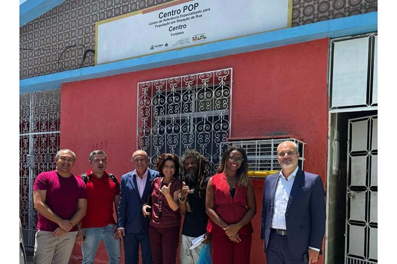 Audiodescrição: Uma foto mostra sete pessoas, quatro homens e três mulheres, posando em frente a um prédio de cor vermelha com uma placa indicando "Centro POP". O prédio tem uma porta e janelas com grades. Todos estão sorrindo para a câmera.
