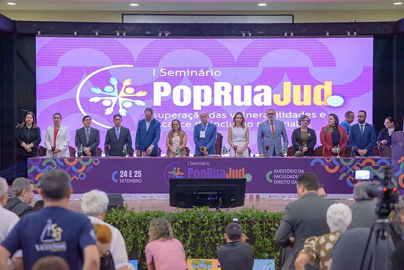 ma mesa de autoridades, com 13 pessoas formalmente vestidas, dispostas em semicírculo, está em frente a uma tela roxa iluminada que exibe o logo "I Seminário PopRuaJud". Há um público em cadeiras à frente, desfocado, e algumas plantas decorativas na base da mesa.