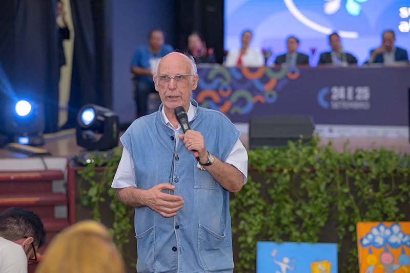 O Padre Júlio Lancellotti, um senhor branco e calvo, de óculos, veste uma camisa branca de manga curta e um colete jeans claro. Ele está no centro, segurando um microfone com a mão direita e gesticulando com a esquerda, falando ao público. Ao fundo e fora de foco, vê-se a mesa de autoridades e um telão com a data do evento (24 e 25 de setembro).