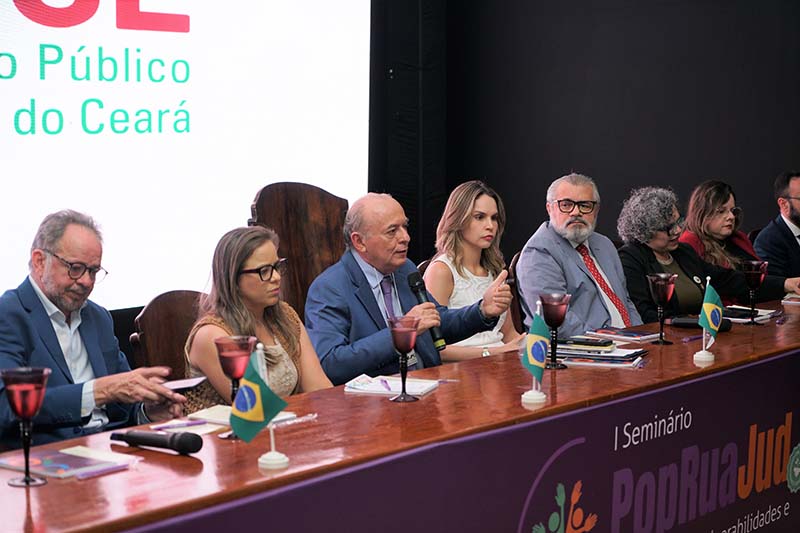 Close-up de sete autoridades sentadas à mesa, com enfoque no centro. O homem no centro (Desembargador José Antônio Parente da Silva, no contexto do evento) está falando, gesticulando com a mão direita e olhando para o lado esquerdo. Copos de vinho e bandeiras do Brasil estão sobre a mesa de madeira, e ao fundo, um telão branco exibe um texto cortado.