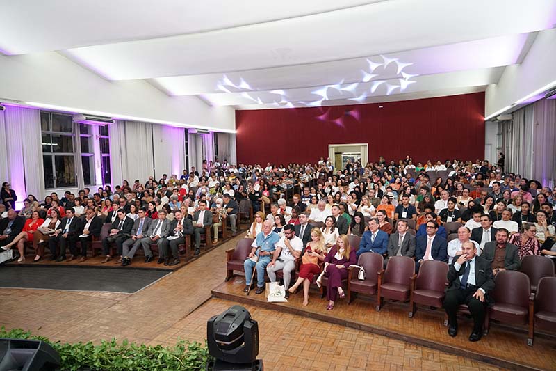 Vista panorâmica do auditório completamente lotado com uma grande plateia sentada em cadeiras estofadas marrons. O público, em sua maioria formalmente vestido, preenche as laterais e o centro do salão, que tem piso de madeira. O fundo da sala é uma parede em tom bordô com uma porta central.