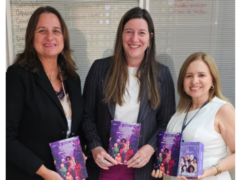 Três mulheres sorriem para a câmera, cada uma segurando um livro de capa roxa com ilustrações coloridas de mulheres. A mulher do centro veste um terno listrado e as das laterais, blazers. Ao fundo, um quadro branco com anotações em caneta.