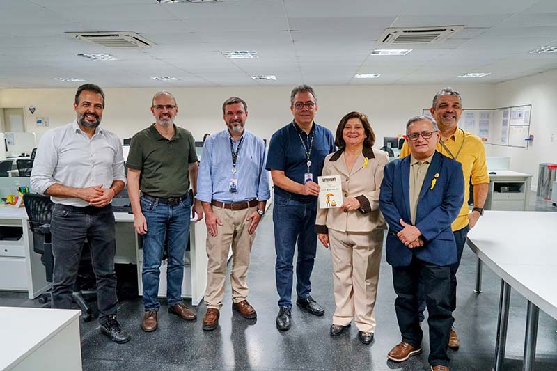 A foto em ambiente interno mostra sete pessoas, em pé, lado a lado. A maioria é de homens. Ao centro, uma mulher de tailleur bege, usa um broche de fita amarela e segura uma cartilha.