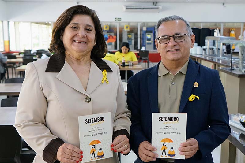 Uma foto em um refeitório, com duas pessoas em pé, sorrindo para a câmera. Ao lado direito, um homem de blazer azul segura uma cartilha. Ao lado esquerdo, uma mulher de tailleur bege segura uma cartilha. Ambos usam broche de fita amarela.