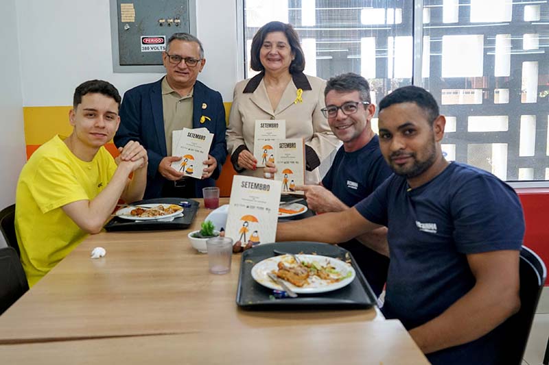 Foto em ambiente de refeitório, com cinco pessoas sorrindo ao redor de uma mesa. Na frente, à esquerda e à direita, dois homens de camiseta seguram um livro de capa branca, assim como as duas pessoas do meio, um homem e uma mulher. A pessoa atrás, na esquerda, não segura nada.