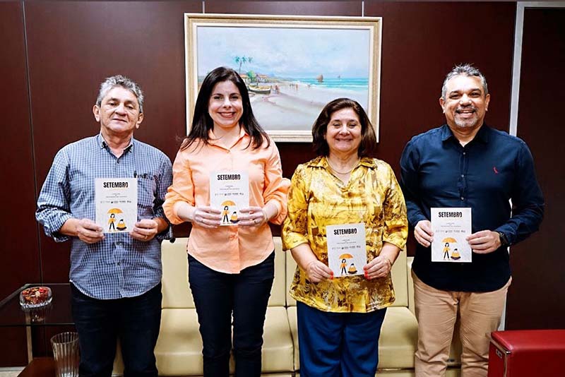 Quatro pessoas, duas mulheres e dois homens, posam para uma foto em um escritório. Eles seguram a "Cartilha de Prevenção ao Suicídio".