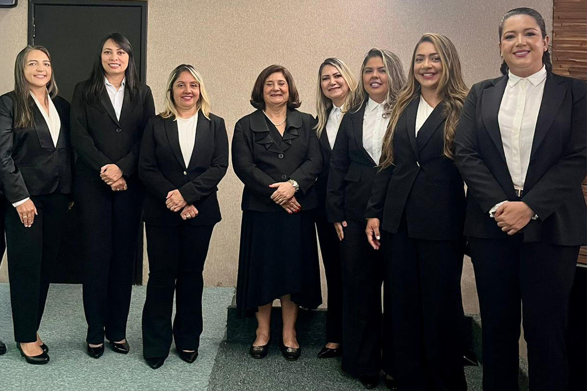 Oito mulheres estão em pé, posando juntas para uma foto em um ambiente interno com paredes claras. Todas vestem trajes sociais escuros, predominantemente blazers e calças pretas. A mulher ao centro, um pouco mais baixa, usa um blazer de botões e saia, enquanto as outras a rodeiam com sorrisos discretos. A iluminação é suave e o grupo parece coeso e profissional.