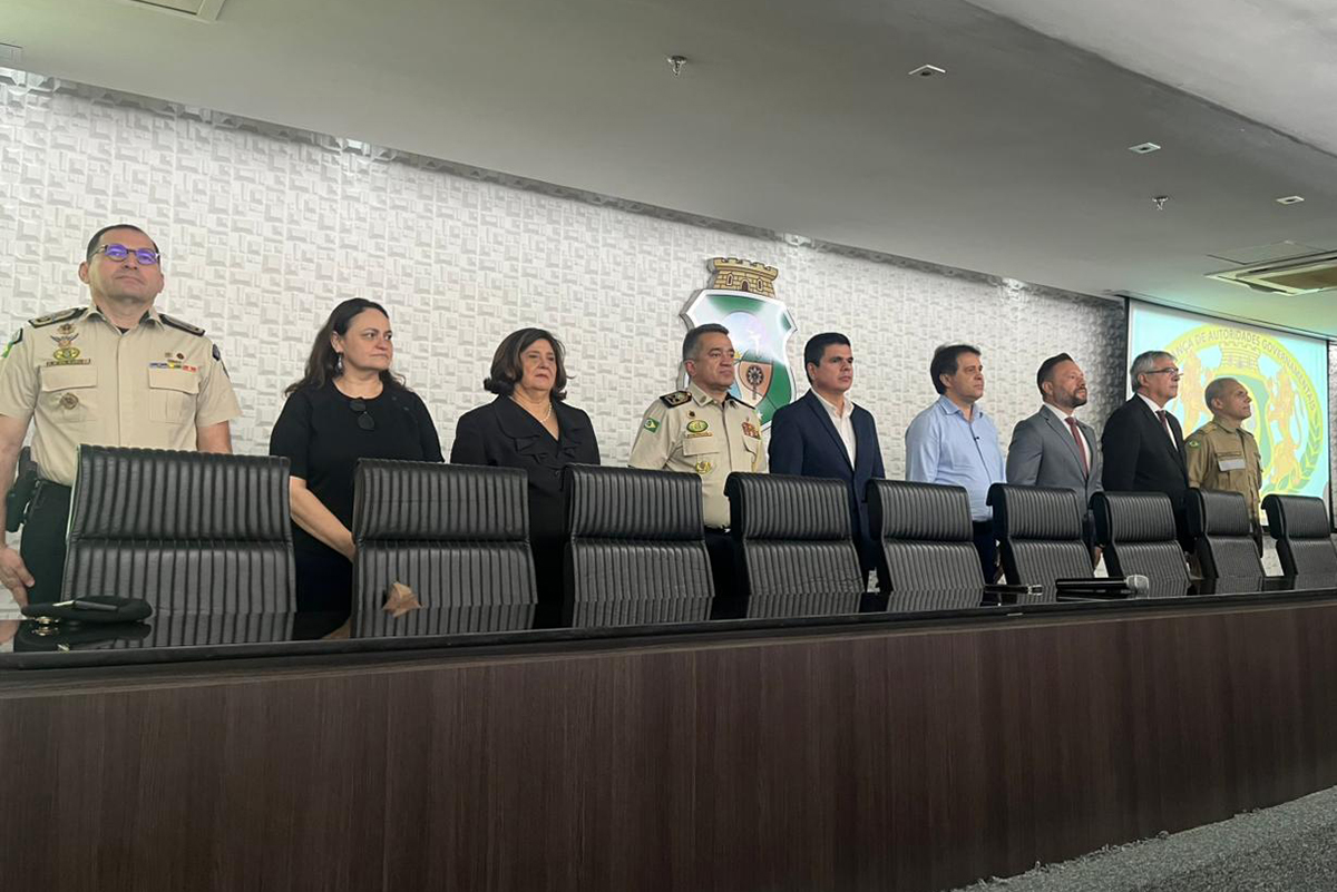 Oito pessoas, sendo seis homens e duas mulheres, estão em pé atrás de uma longa mesa de madeira escura com cadeiras pretas. A maioria veste trajes formais, como ternos e blazers, e dois homens usam fardas da polícia militar. Ao fundo, uma parede texturizada branca exibe um brasão e parte de uma tela de projeção. Eles estão perfilados, olhando para a frente, em uma aparente cerimônia ou reunião.