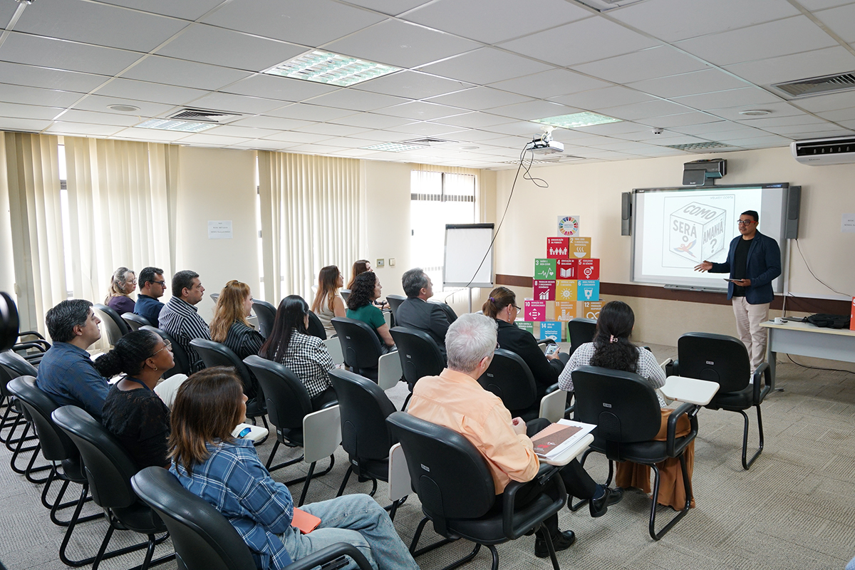Em uma sala de conferências com teto de placas brancas e iluminação embutida, um palestrante gesticula em frente a uma tela de projeção. A plateia, composta por homens e mulheres de diversas idades, está sentada em cadeiras pretas e observa atentamente a apresentação. Ao lado da tela, há cubos coloridos dos ODS empilhados. A projeção mostra a pergunta "COMO SERÁ O AMANHÃ?".