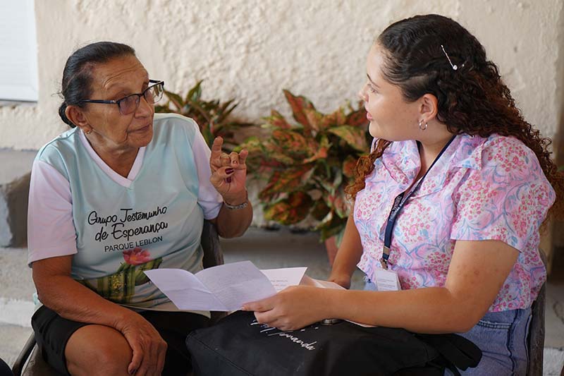 Uma jovem e uma senhora idosa estão sentadas e engajadas em uma conversa. A idosa, vestindo a camisa do "Grupo Testemunho de Esperança", gesticula com a mão enquanto fala. A jovem, com cabelo cacheado e vestindo uma blusa estampada, ouve atentamente, segurando papéis e com uma bolsa no colo.