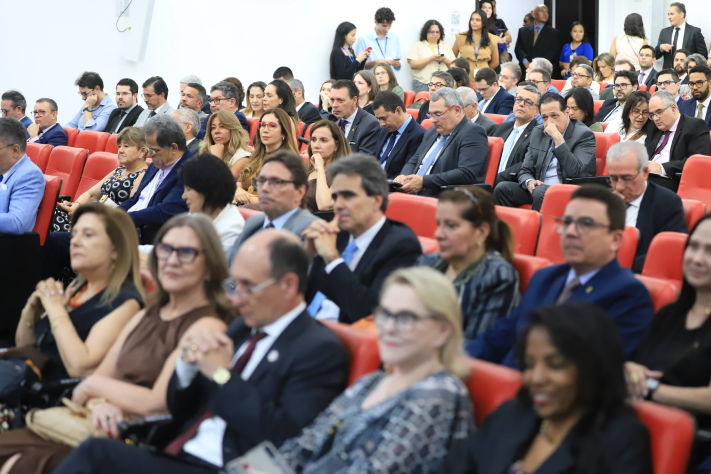 Uma ampla foto mostra uma plateia lotada de um auditório, com diversas pessoas sentadas em poltronas vermelhas, atentas à frente. No primeiro plano, à direita, está a Desembargadora Adenir Carruesco (cabelos loiros e óculos) ao lado da Desembargadora Laís Helena Jaeger Nicotti (de blusa escura). Outras autoridades, homens e mulheres, ocupam as fileiras de trás.