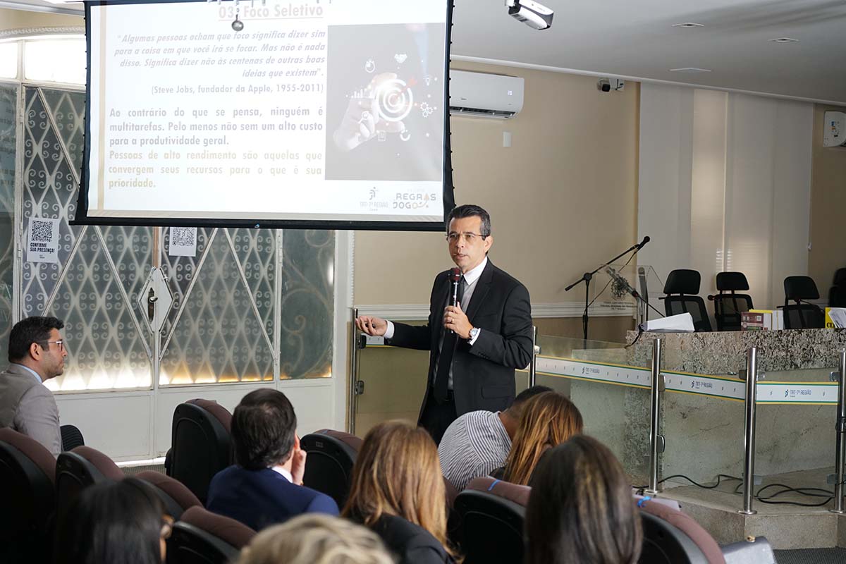 Um homem em terno escuro, com microfone na mão, gesticula em frente a uma tela de projeção, onde há um slide com uma citação de Steve Jobs. Ele está apresentando uma palestra em um auditório fechado, com algumas pessoas sentadas na plateia.