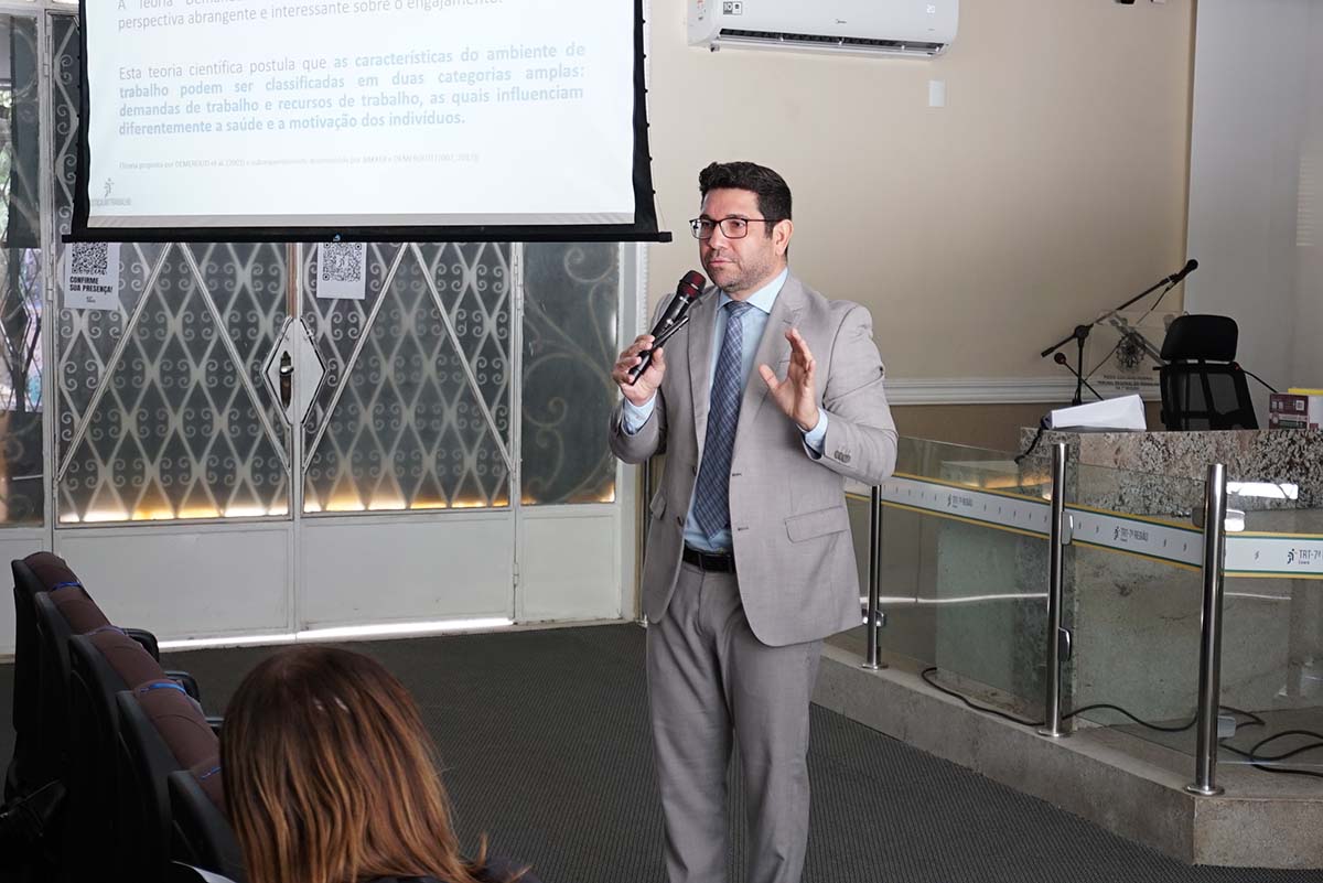 Um homem em terno cinza claro, com gravata azul, segura um microfone e gesticula com as duas mãos abertas, enquanto fala no palco de um auditório. Atrás dele, uma tela de projeção exibe um slide com texto científico sobre ambiente de trabalho.