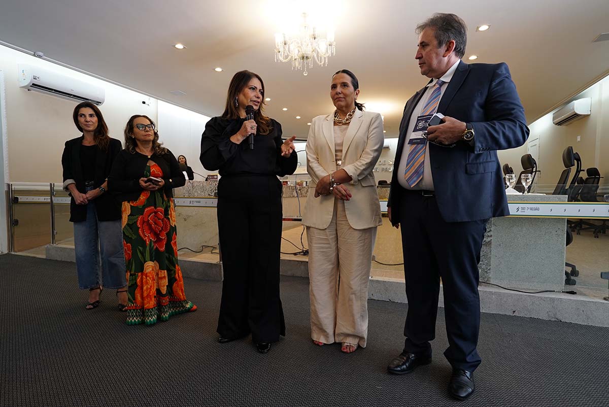 Quatro pessoas, sendo três mulheres e um homem, estão de pé na frente do auditório. A Desembargadora Ruth Barbosa Sampaio, vestindo preto, segura um microfone e gesticula. Ao lado dela, o Diretor da Ejud-7, Paulo Régis Machado Botelho, a observa, acompanhado por outras integrantes do CONEMATRA.