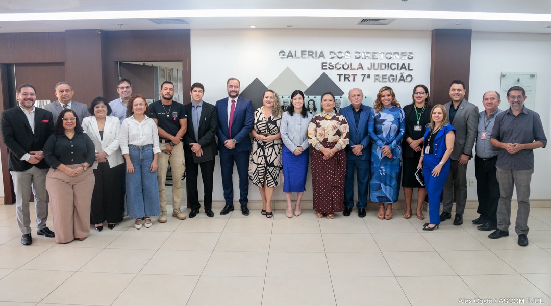 Em uma sala iluminada, um grupo diverso de vinte pessoas, incluindo homens e mulheres de várias etnias e idades, está posicionado em fila para uma foto. Ao fundo, um painel em tons de cinza exibe "GALERIA DOS DIRETORES ESCOLA JUDICIAL TRT 7ª REGIÃO". Todos sorriem, vestidos com trajes formais e casuais.