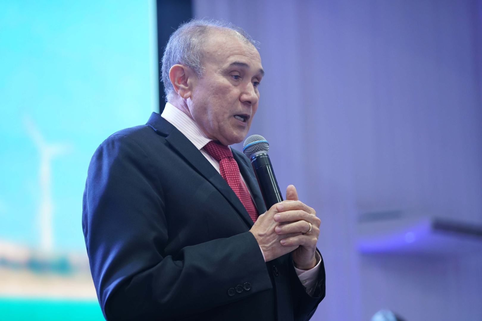 Um homem idoso de pele clara e calvície parcial, vestido com um terno escuro, camisa listrada em rosa e gravata vermelha texturizada, está de perfil parcial à esquerda, segurando um microfone com as duas mãos. Ele parece estar falando com seriedade, com a boca aberta. O fundo é iluminado em azul claro, com uma sutil imagem verde e branca, talvez de uma turbina eólica, na parte inferior esquerda.