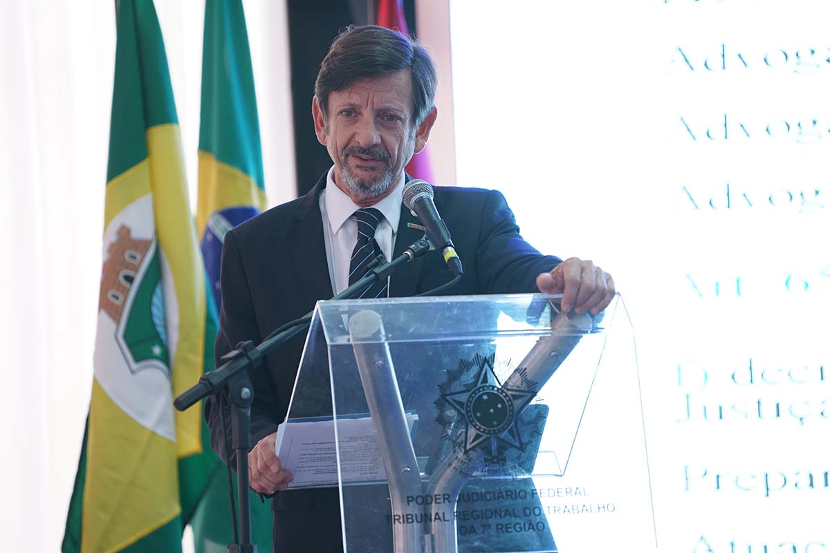 Um homem de pele clara e barba grisalha, vestindo terno e gravata, aparece da cintura para cima. Ele segura com a mão direita a lateral de um púlpito de acrílico que exibe o logo do Poder Judiciário Federal - Tribunal Regional do Trabalho da 7ª Região. Ao fundo, parcialmente visíveis, estão as bandeiras do Brasil, Ceará e a do TRT-7.