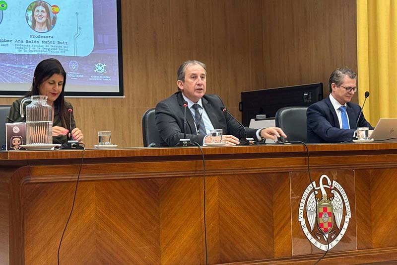 Três pessoas (duas mulheres e um homem no centro) estão sentadas em uma mesa de madeira escura com microfones e copos d'água, durante uma conferência. Ao fundo, um telão parcial mostra o nome "Professora Ana Belén Muñoz Ruiz". O homem do centro está falando e gesticulando.