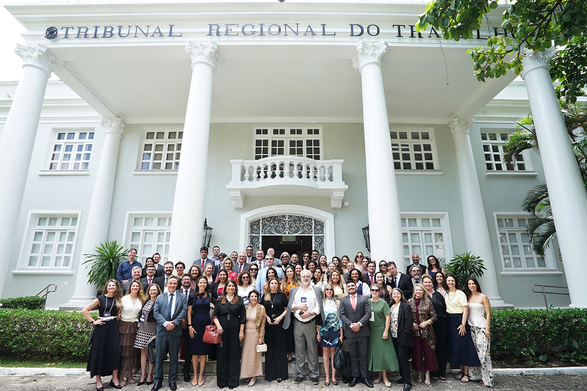 Uma foto em plano geral mostra um grupo grande e diversificado de pessoas, homens e mulheres, de pé e sentados, em frente a um edifício imponente e clássico. O prédio tem uma fachada cinza-clara, com colunas brancas de estilo grego sustentando uma varanda, e janelas altas. O letreiro no topo do edifício diz TRIBUNAL REGIONAL DO TRABALHO. O grupo está bem vestido, a maioria com trajes sociais ou esporte fino, posando para a foto em degraus e no gramado à frente da construção.
